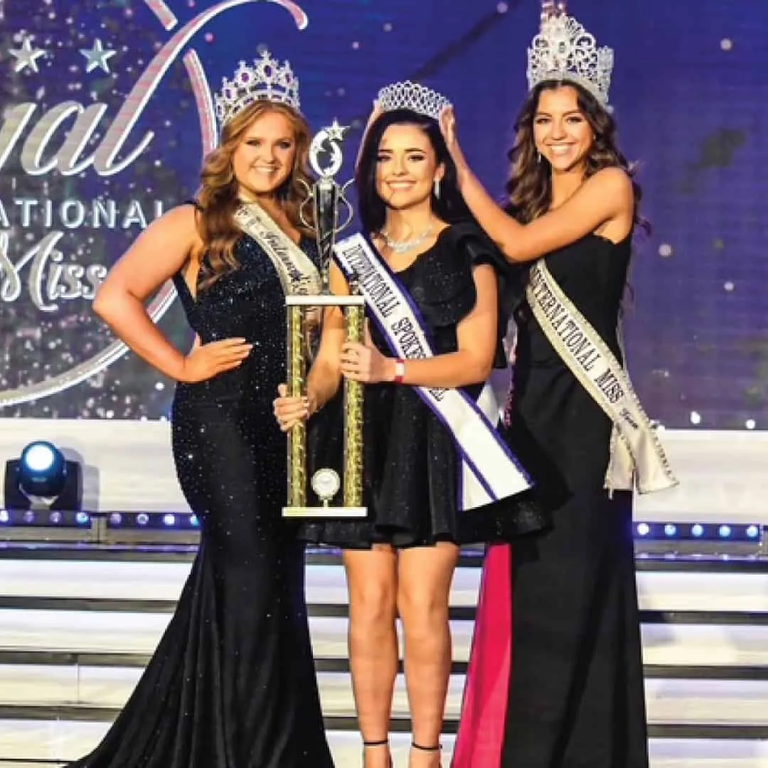 Three crowned Queens in black gowns with a trophy