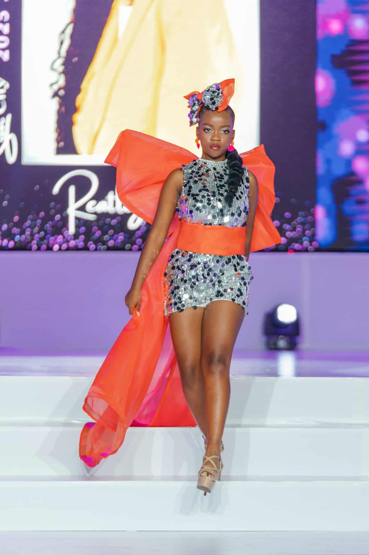 Bright Miss SA pageant contestant in glamorous sequin gown with bold orange accents and dramatic accessories at Royal International Miss SA event.