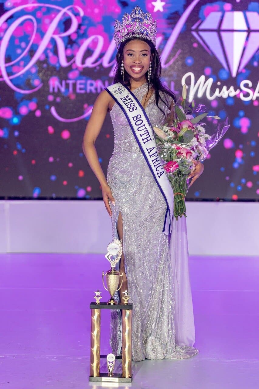 Elegant young woman crowned Miss SA, wearing a sparkling gown and sash, holding a bouquet of flowers, standing on stage at the Royal International Miss SA pageant.