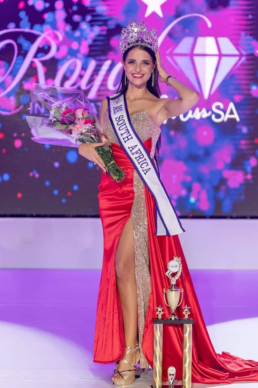 Elegant young woman crowned as Royal International Miss South Africa, wearing a stunning red evening gown, holding a bouquet of flowers and trophy, celebrating her beauty and achievement at the pageant.