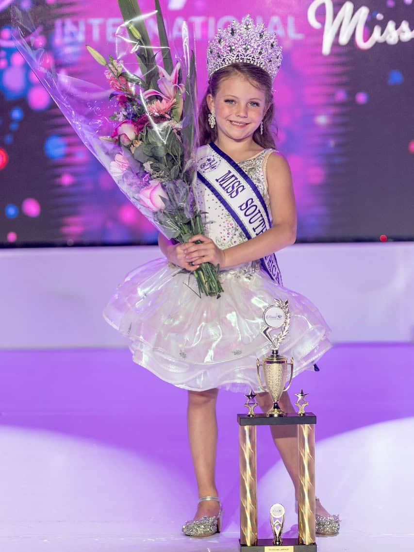 Bright young girl crowned at Royal International Miss SA pageant, holding a bouquet and standing next to her trophy with sash and tiara, showcasing beauty and confidence in this South African beauty contest.