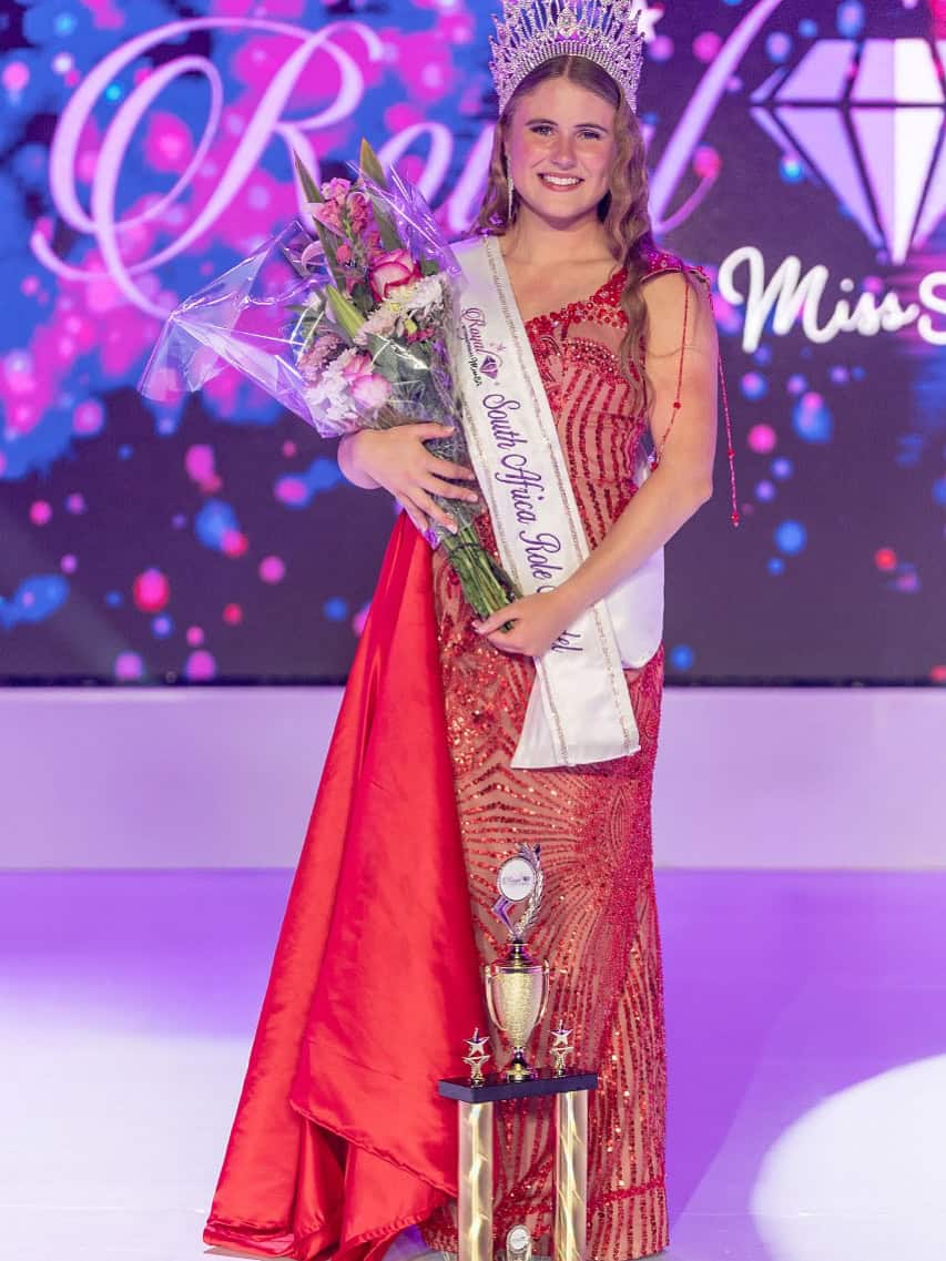 Elegant young woman crowned Miss South Africa at Royal International Miss SA pageant, holding flowers, wearing a red evening gown and sash, celebrating her victory in South Africa's beauty contest.