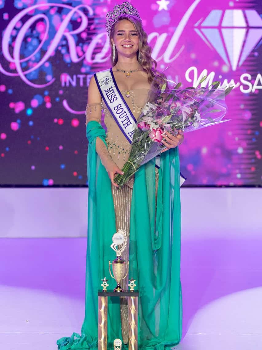 Elegant young woman crowned as Royal International Miss SA, wearing a tiara and sash, holding flowers and standing beside a trophy, celebrating her victory in a beauty pageant event.
